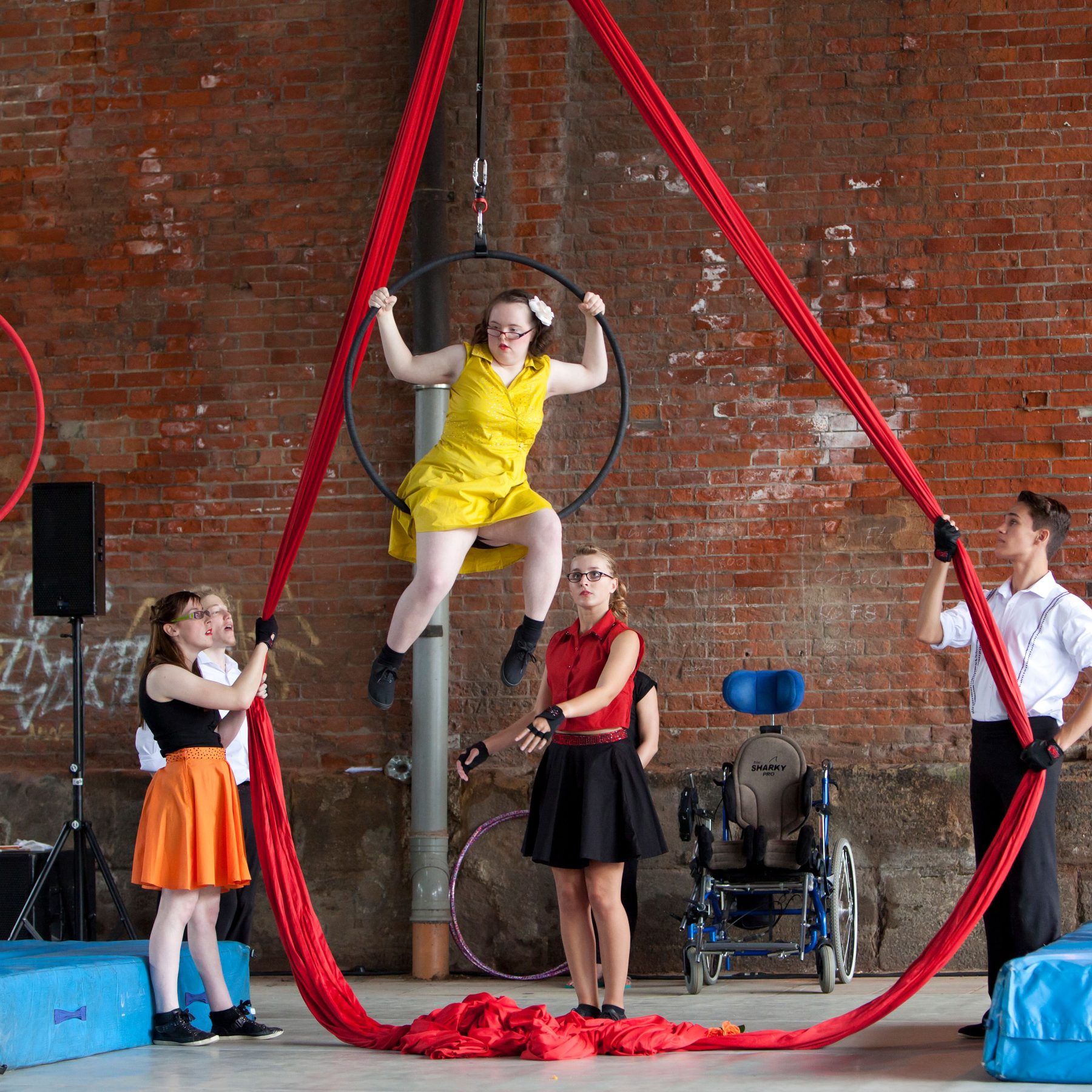 A young integrated group of circus performers wear bright colours as they perform their show. A girl hangs from an aerial hoop whilst two others hold red aerial silks.