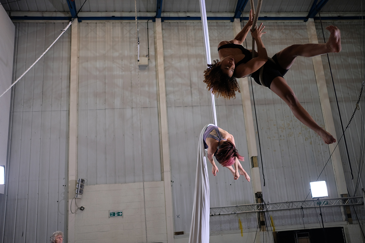 Extraordinary Bodies: Two female aerialists, one black with brown afro hair and the other visually impaired with red hair swing from the ceiling, their limbs flying in all directions as the dance on their aerial equipment.