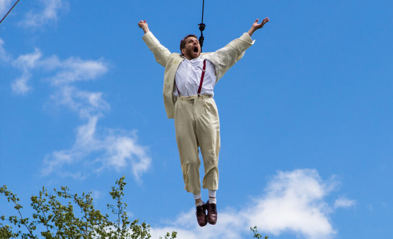 Jamie flies in the air on an aerial harness, his arms outstretched with an ecstatic expression on his face, a bright blue sky behind him.