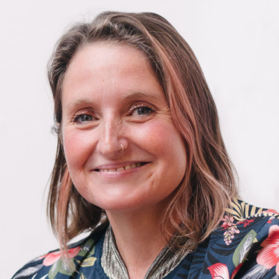 Headshot of Claire: a fifty year old white woman with shoulder-length brown hair, a pierced nose and pierced ears, green eyes, a rosy complexion, a big smile, and her own teeth (so far).