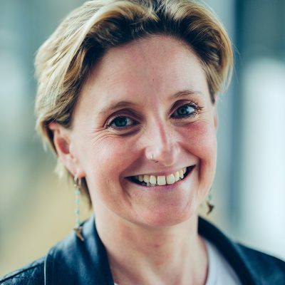 Headshot of Claire: a fifty year old white woman with shoulder-length brown hair, a pierced nose and pierced ears, green eyes, a rosy complexion, a big smile, and her own teeth (so far).