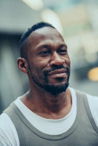 Headshot of David, a Black man with a stubble beard, shirt black hair and a soft smile> He wears a beige tank top over a white T-shirt.