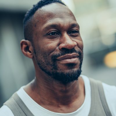 Headshot of David, a Black man with a stubble beard, shirt black hair and a soft smile> He wears a beige tank top over a white T-shirt.