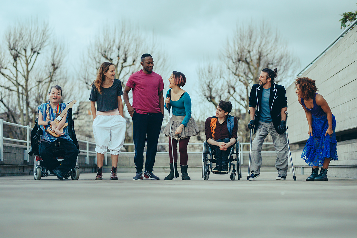 Seven artists line up outside standing on grey concrete. They wear colourful clothes and look towards each other laughing. Two of the cast are in wheelchairs, one holds a stick and one holds crutches. They are a mix of male female, black, white and mixed race, disabled non-disabled.