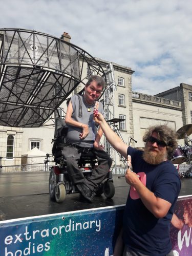 John Kelly sits on an outdoor stage in his wheelchair whilst Dave the production manager passes him an Ice Lolly covered in colourful sprinkles