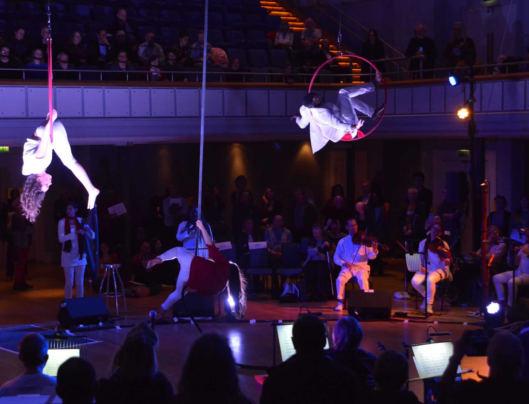 Wide shot of Birmingham Town Hall, the musicians seated in a large circle around the room, creating a centre stage for the aerial artists. One girl suspended upside down in a red hoop, another man perches with his legs high, sat in another red hoop. A second girl is flowing down a rope, also suspended from the ceiling