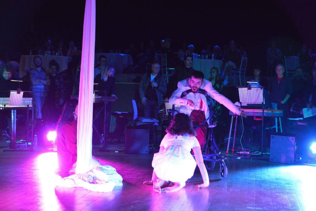 Dave Young moves across the stage in his walker, a girl crouches on the floor in front of him. Another girl sits at the bottom of the silks to their left. The corner of the stage is illuminated with pink light.