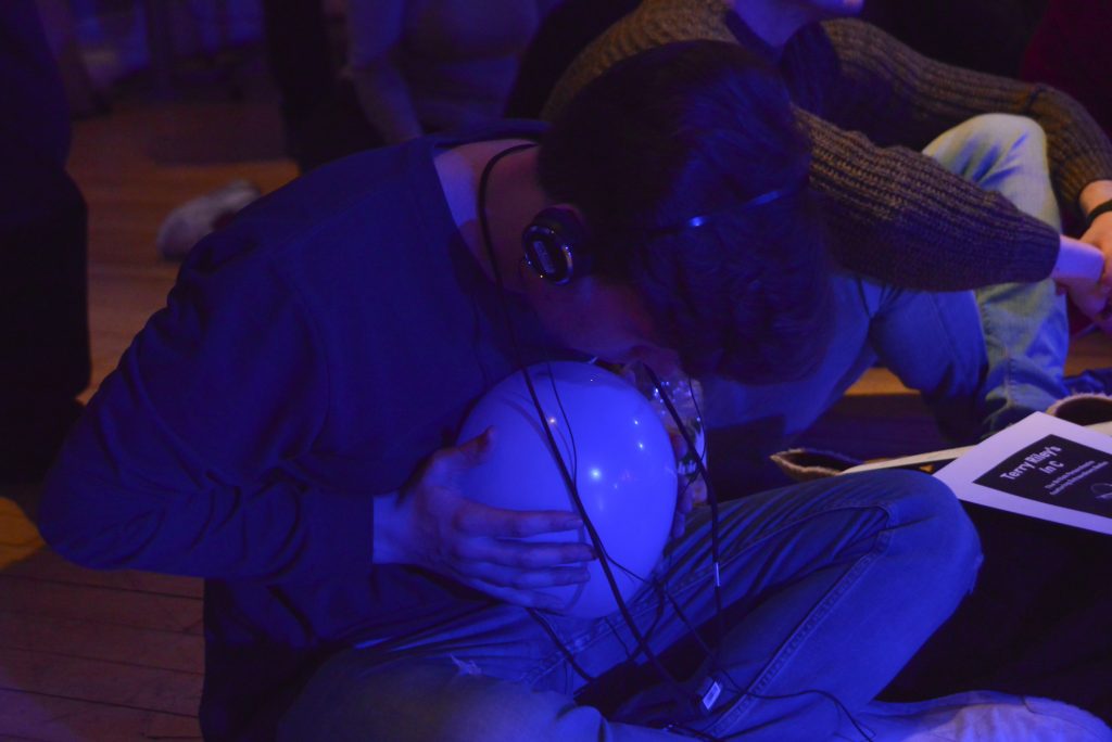 An audience member is seated, cross legged, holding a small inflated balloon. They wear headphones and curl up over the balloon to feel the reverberations from the live music. A beautiful way to experience a live performance.