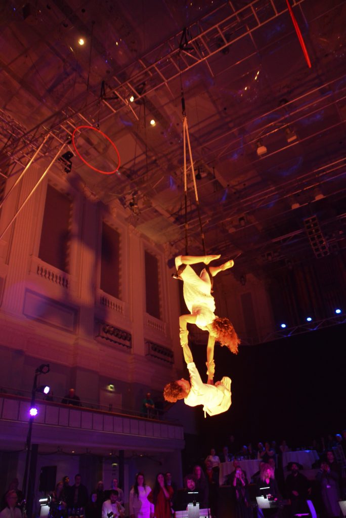 An aerialist is suspended from a trapeze from her legs, the large ceiling of Birmingham Town Hall towers up above. She holds a man by the strength of his arms, dangling above the musicians.