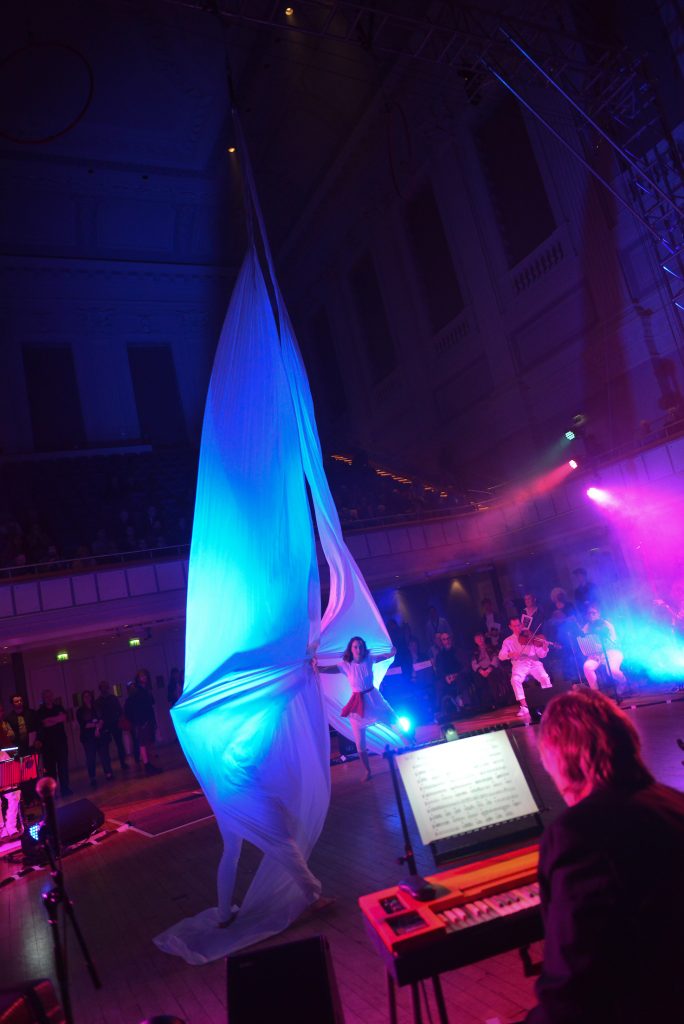 Artists create a spectacle in the centre of the large town hall, as the silks billow into large sails, illuminated by bright blue light. Musicians of the Paraorchestra surround them in a large circle.