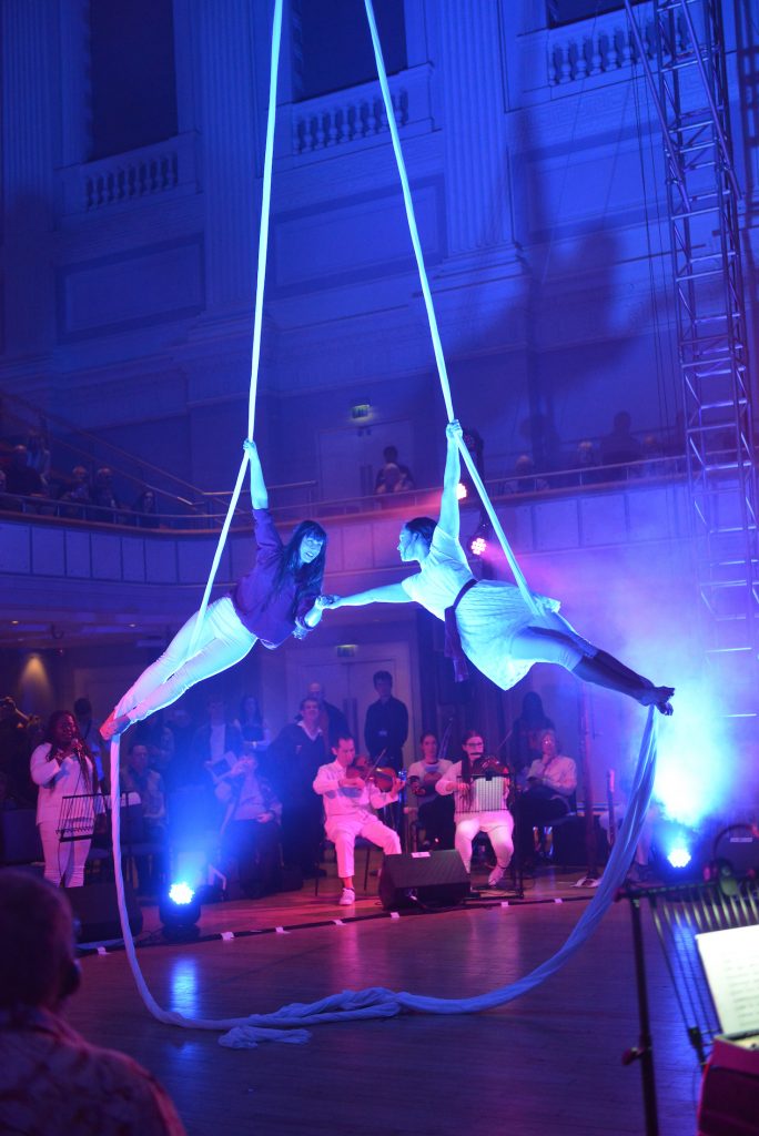 Two female aerialists suspended from silks in the centre of the floor stage. Their arms outreached and hands touch.