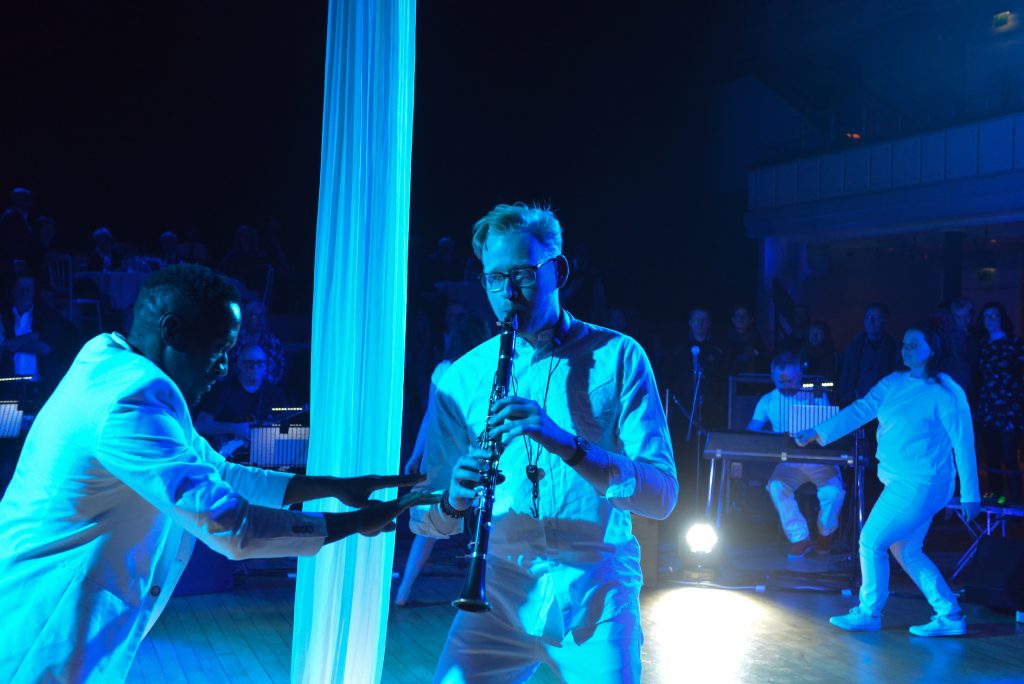 A man playing the clarinet walks across the central stage space, silks illuminated in bright blue light behind him. Another man moves around him to the rhythm with arms outstretched.