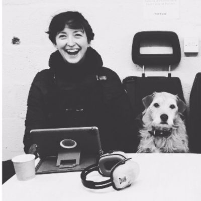 Photograph of Georgina, laughing to camera. She's a white woman, with short dark hair. She's sat next to a small scruffy light haired dog.