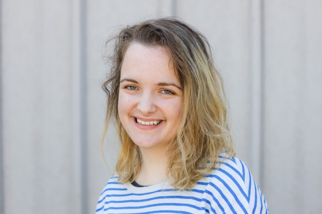 Nikki poses for the camera. She is a young white woman with dirty blonde shoulder length hair. She smiles softly. She is wearing a white and blue striped top.