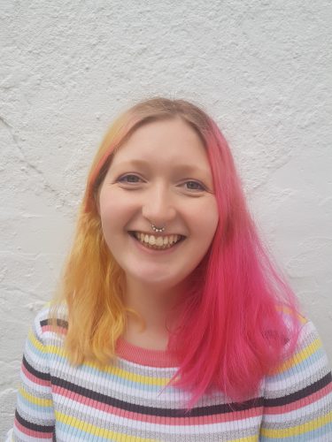 Ellie Stephens, smiling to camera - a young white girl, with long shoulder length hair - one side yellow blonde and the other side dyed bright pink. Ellie smiles. Her septum nose piercing is just visible.