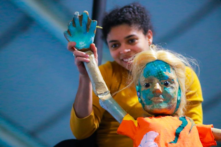 A young mixed-race woman is smiling and holding up a puppet of a girl with blond hair, an orange top and fish-like turquoise scales of her face.