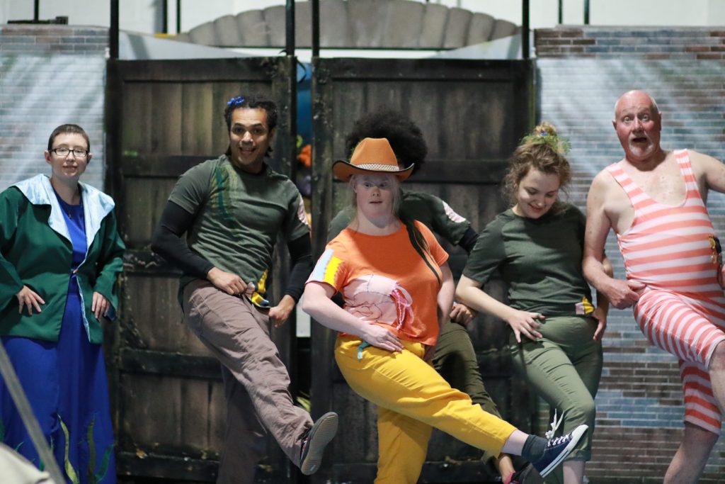 The cast line dance with Helen Cherry as Flo in the middle wearing a cowboy hat. 