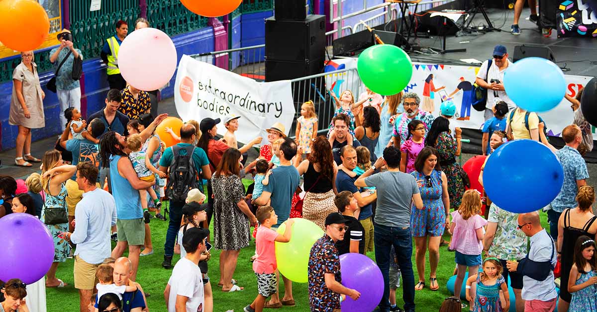 A crowd of people at an outdoors event. Colourful giant balloons flying in the air. A large white banner with a light red circle and black logo that reads: "Extraordinary Bodies".