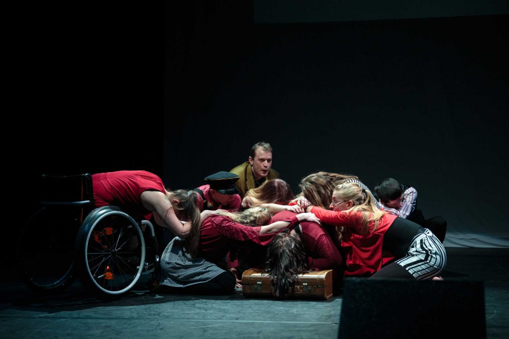 A group of young disabled and non-disabled people grouped together in the centre of a dimly lit stage. They are all sitting or bending their bodies towards the ground, holding onto each other.