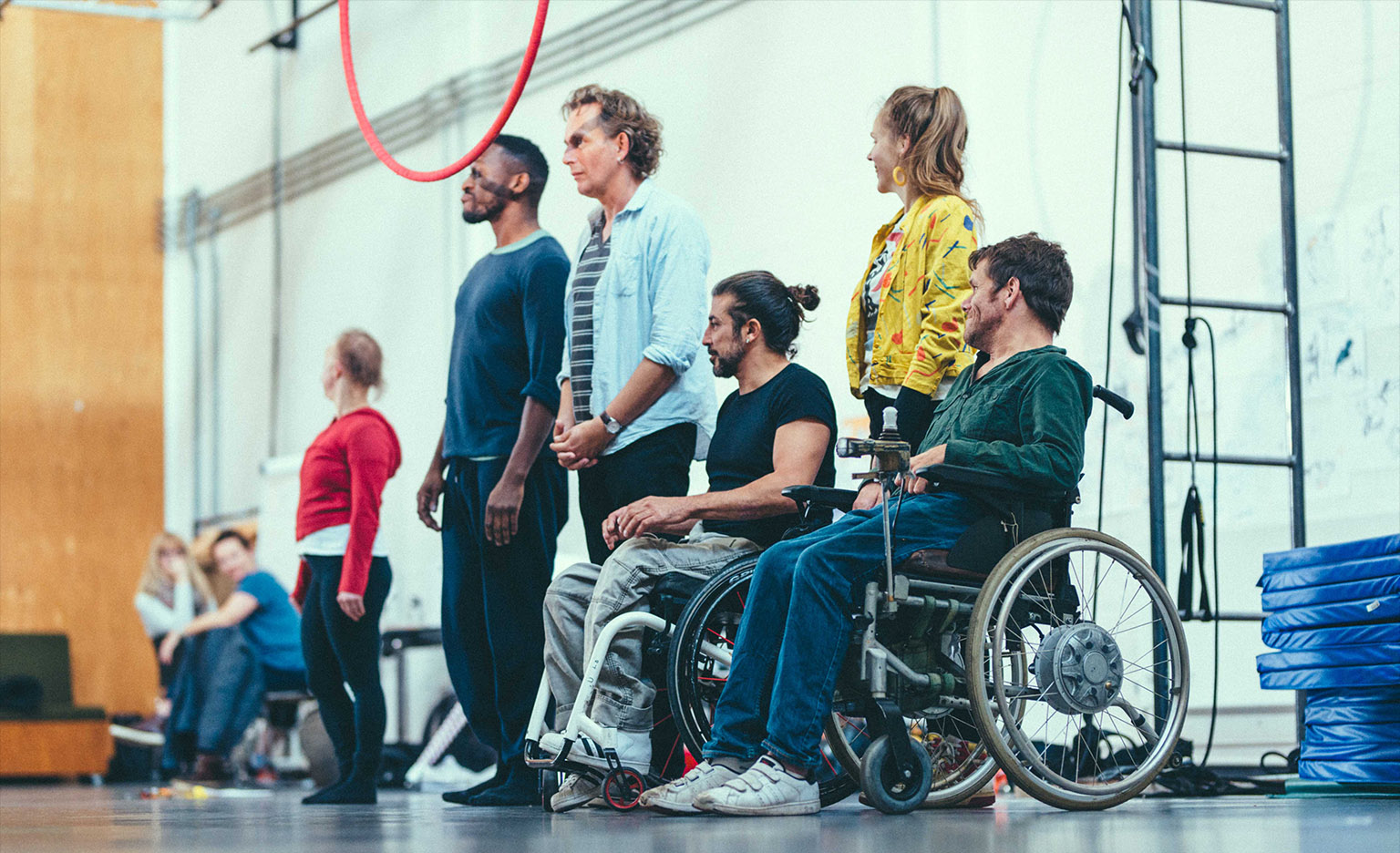 A group of disabled and non-disabled, black and white artists stand and sit in wheelchairs in a rehearsal space. A circus hoop hands from the ceiling and crash mats are visible on the side.