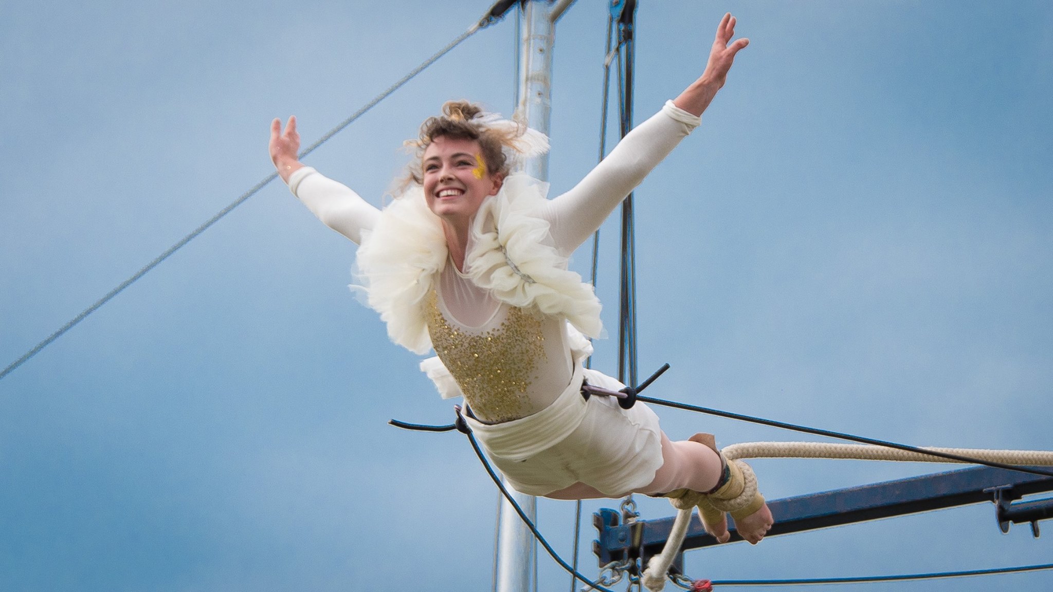 Tilly flies through the air, smiling brightly, her feet tied to a circus rope. Outdoors rigging and a grey sky behind her.