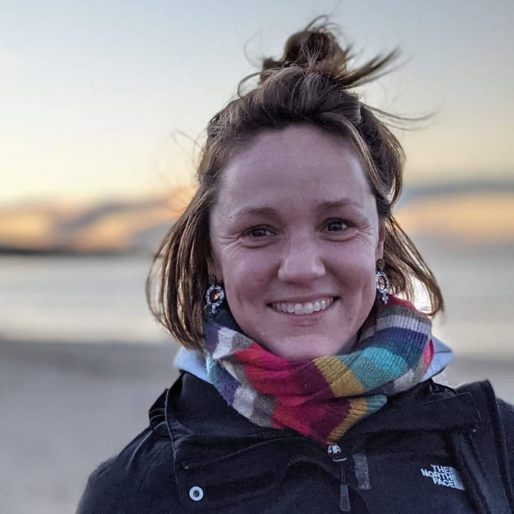 Jodie - shoulder-length brown hair, a bright smile, a colourful scarf. She is standing on a beach, with a sunset and sea in the background.