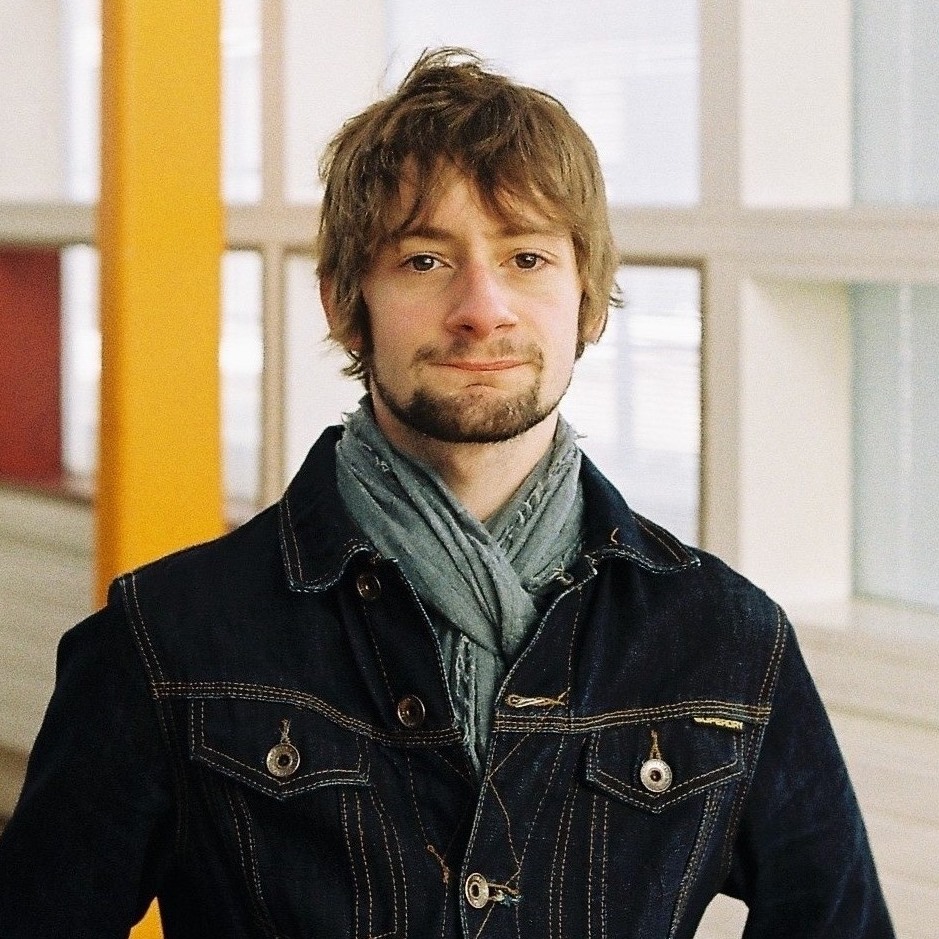 Oliver poses for the camera. He has mice brown hair that falls over his ears down to his cheekbones and a short beard. He wears a grey-blue scarf and a dark-blue denim jacket.