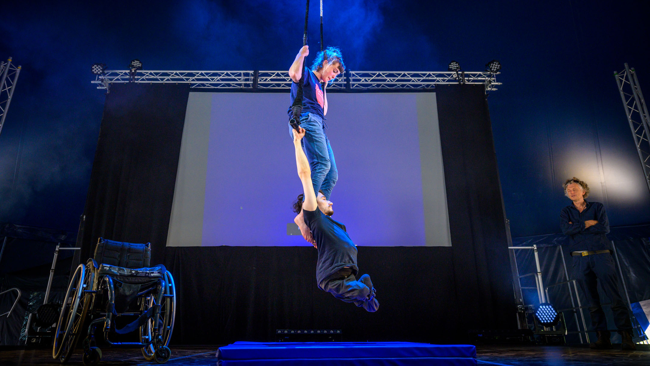 Indoors circus stage. A white woman in her twenties sits on a trapeze, her hands holding the ropes on each side. A white man in his twenties and wheelchair user holds himself up in the air below her by holding onto the trapeze bar. Her legs extend down to his upper back, her feet by his shoulders.