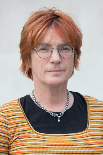 Headshot of Abi, a white woman with short hair dyed orange, round and thin-rimmed glasses. She has a nose ring and wears a thick chain around her neck.