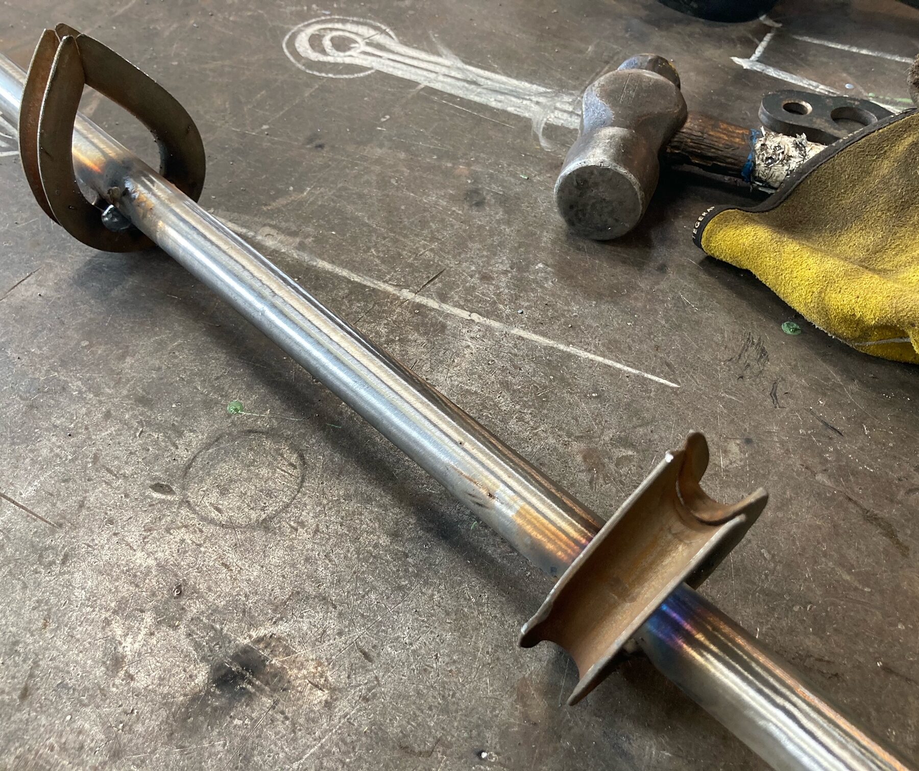 A metal trapeze lies on a metal table. In the background, a hammer and a yellow cloth.