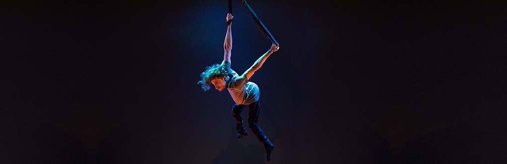 A white man in his twenties in the air above the the stage by holding onto black straps. He wears a grey T-shirt and black trousers. His thin legs dangle below him as he flies, looking strong.