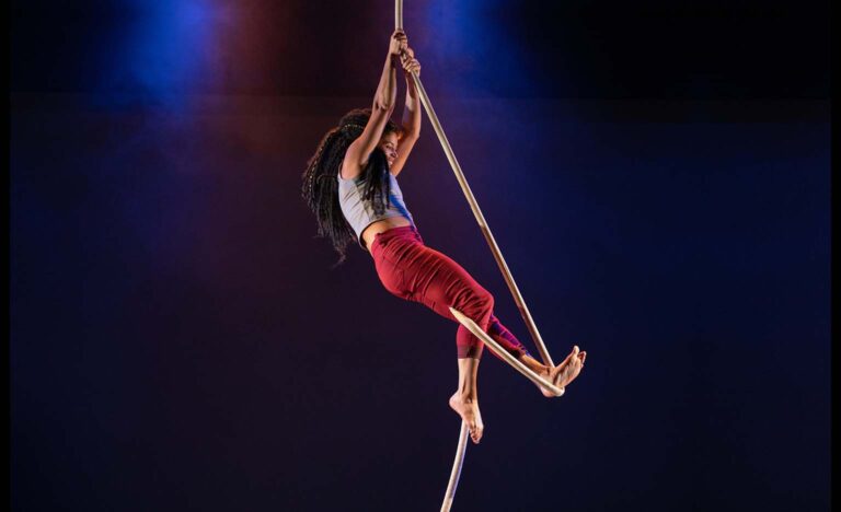 A short, muscular brown skinned woman hangs onto a thick rope with both hands, suspended in the air. The rope is held taut under her left foot and up behind her right knee. She wears a short sleeved grey shirt and tight red trousers. She wears her long, thick, dark hair half up half down.