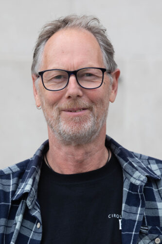 Headshot of James: A middle-aged man with grey hair, a white stubble beard, black-framed glasses. He is wearing a blue and white checked shirt and is smiling softly at the camera.