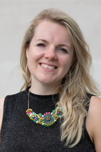 Headshot of Kate, a white woman with long blonde hair and blue eyes. She wears a large necklace made with colourful beads and hung of a thin golden chain, and a sparkly black top.