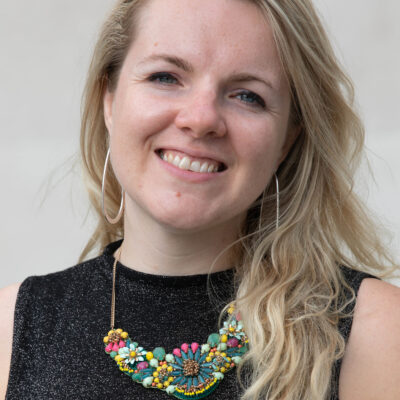 Headshot of Kate, a white woman with long blonde hair and blue eyes. She wears a large necklace made with colourful beads and hung of a thin golden chain, and a sparkly black top.