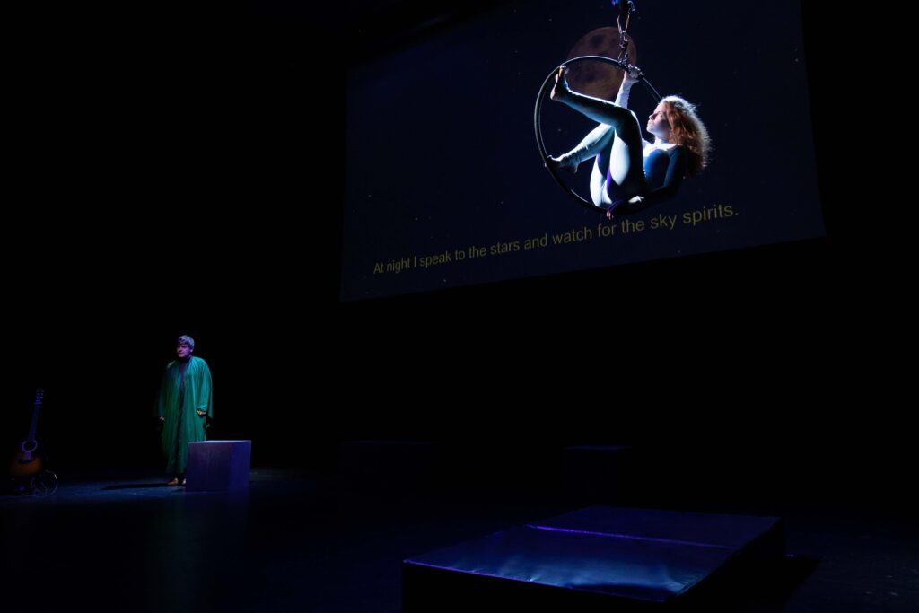 A dimly-lit stage, with soft, deep blue light on two performers. The first one is in the foreground, sitting delicately on a circus hoop that holds them up in the air. They are female-presenting, with long golden hair and a light blue lycra one piece. In the background, a male-presenting performer is dressed like a priest. They stand, looking ahead of them towards the audience.