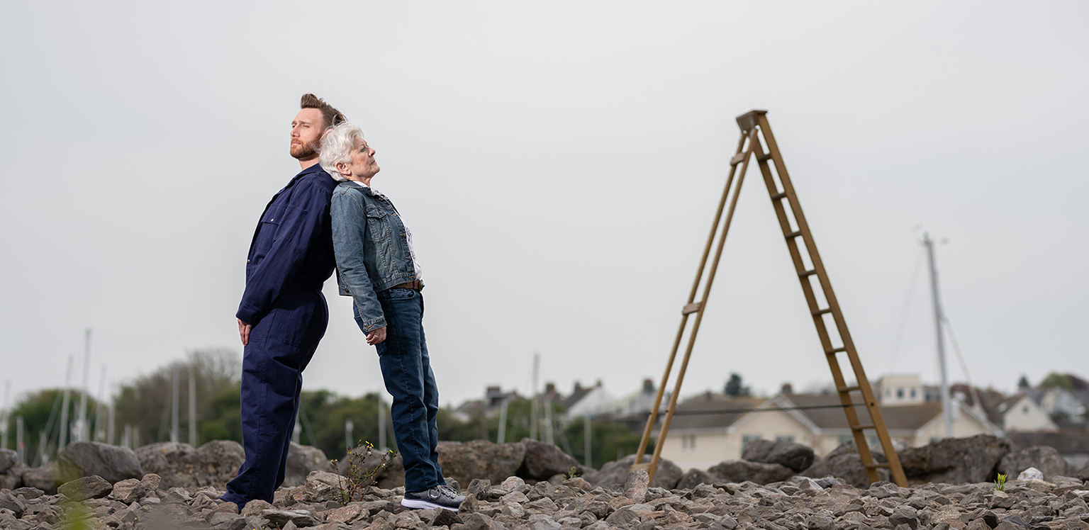 An open area with large pieces of ragged rocks on the ground and a house with trees in the background. Two performers are leaning against each other, back to back, in parallel to the wooden ladder that is stood beside them. The performer to the left is a white man with blonde hair that is slicked back and a ginger beard. He wears a navy blue boilersuit over a white T-shirt. The performer to the right is a white woman in her sixties. She has short white hair, blue eyes and wears a denim jacket with jeans. They are both looking wistfully into the distance.