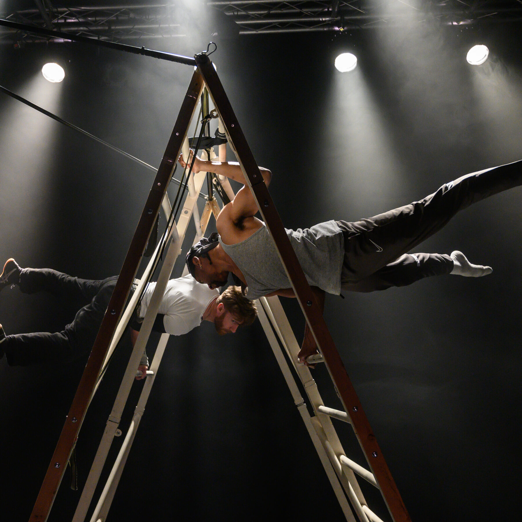 Two performers gracefully balancing betwixt a ladder. Their bodies are parallel to the ground as their heads face each other’s and their legs spread outwards on either side of the ladder. The first performer is a young black man wearing a black bandana, grey tank top, black joggers and grey socks. The second performer is a young white man sporting a beard wearing a white t-shirt, black jeans and black sneakers.