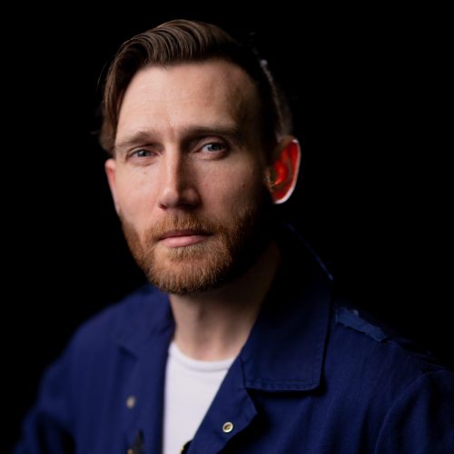 A white man with blonde hair that is slicked back and a stubble beard. He wears a navy blue boilersuit over a white T-shirt.