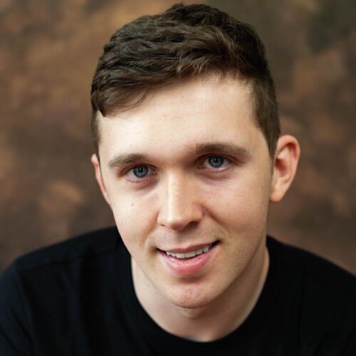 A young white man with short brown wavy hair. He is wearing a black t-shirt and is smiling towards the camera.