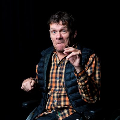 Dark indoor studio. A white man with brown hair, an orange checked shirt and a navy gilet. He is sitting in a wheelchair and looking at the camera with a cheeky expression.