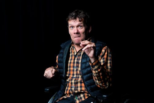 Dark indoor studio. A white man with brown hair, an orange checked shirt and a navy gilet. He is sitting in a wheelchair and looking at the camera with a cheeky expression.