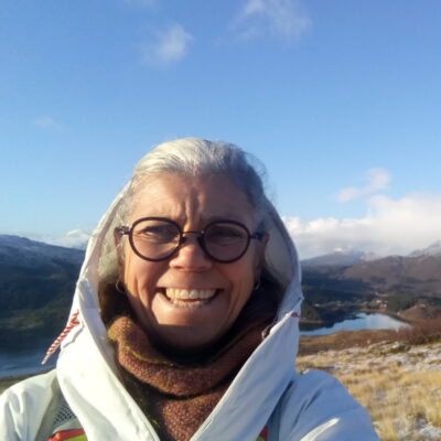 A white woman with white hair wearing glasses and a big white coat with a brown scarf. She is in a frosted landscape with mountains and a lake in the background.