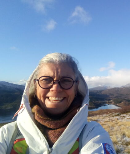 A white woman with white hair wearing glasses and a big white coat with a brown scarf. She is in a frosted landscape with mountains and a lake in the background.