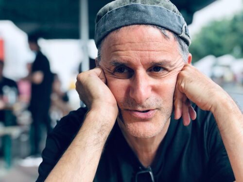 A white man with grey hair wearing a grey cap and a black t-shirt. His hands are resting on his face. He has a captivated expression on his face.