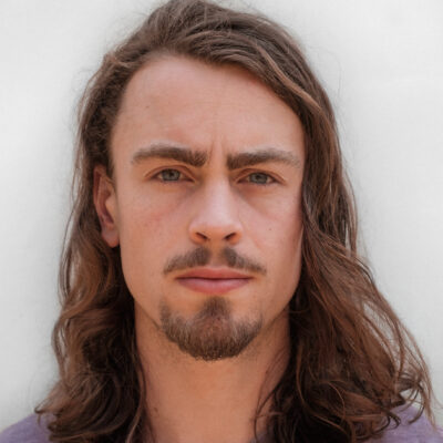 A young white man with long brown wavy hair. He has a goatee and is wearing a purple t-shirt.