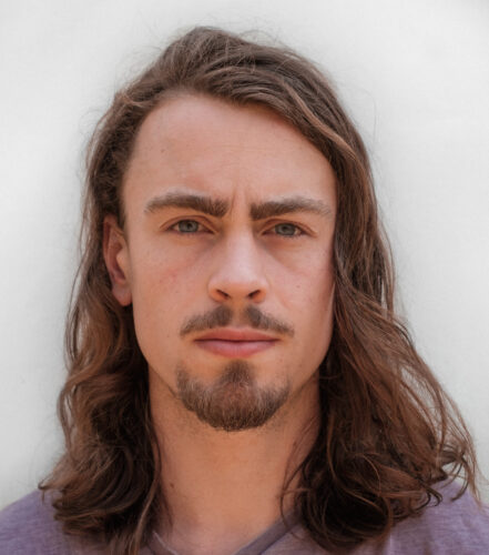 A young white man with long brown wavy hair. He has a goatee and is wearing a purple t-shirt.