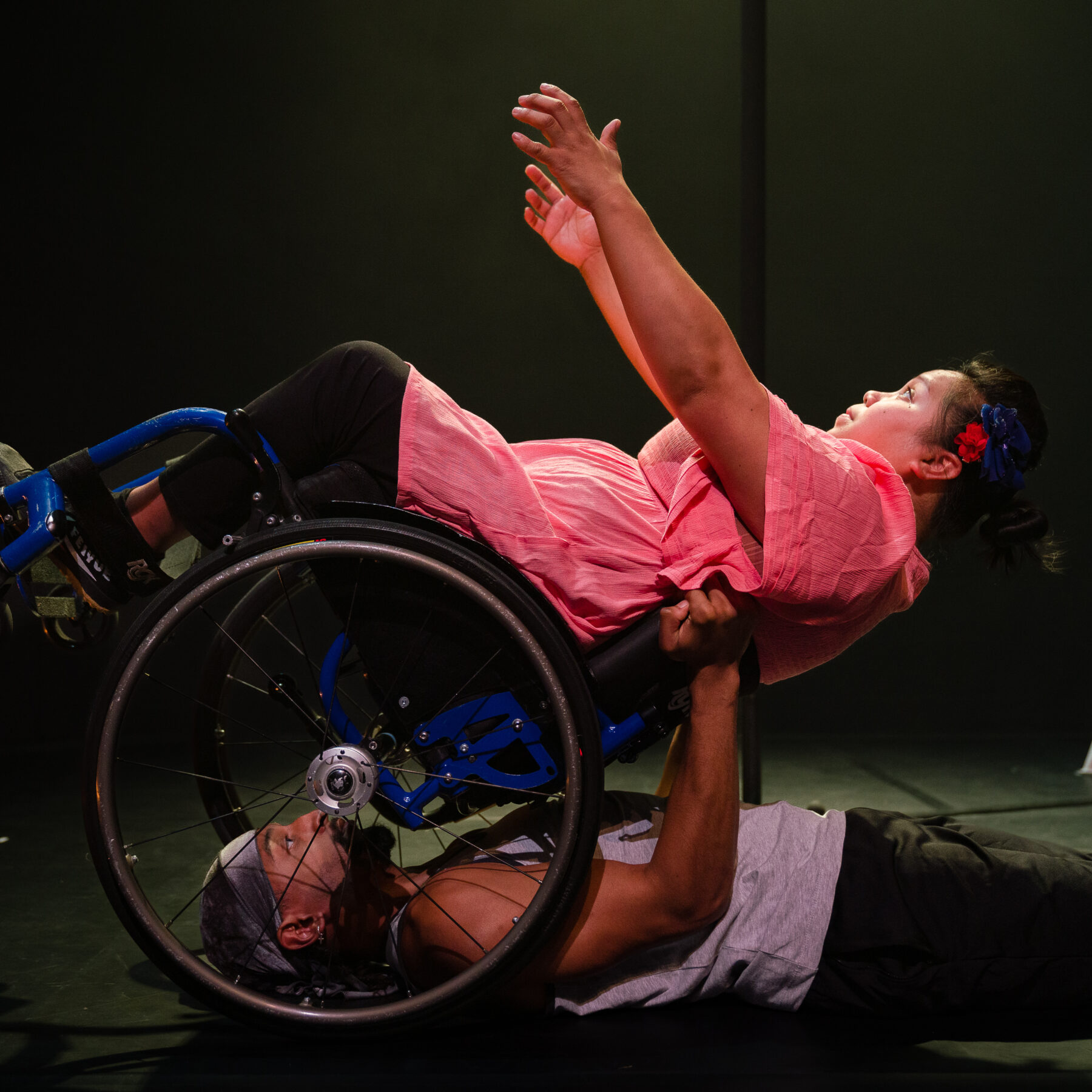 Two performers in a movement of beautiful symmetry, the first performer lying on the ground and holding up the second performer in a wheelchair who is leaning backwards into the hands of the first performer. The first performer is a black young man wearing a black bandana, grey tank top and black joggers. The second performer is a woman in a wheelchair wearing a pink top and leggings.