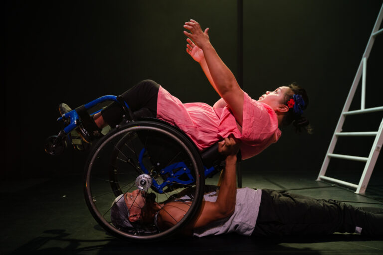 Two performers in a movement of beautiful symmetry, the first performer lying on the ground and holding up the second performer in a wheelchair who is leaning backwards into the hands of the first performer. The first performer is a black young man wearing a black bandana, grey tank top and black joggers. The second performer is a woman in a wheelchair wearing a pink top and leggings.