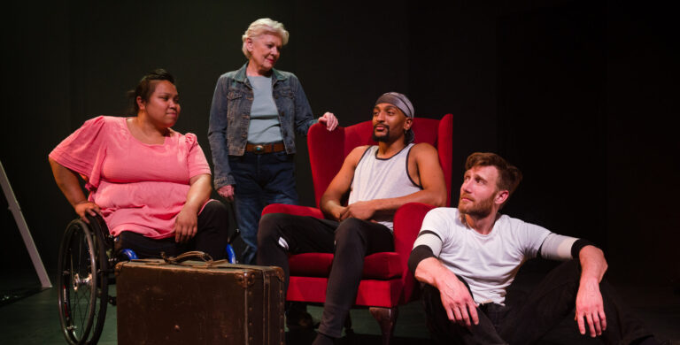 Four performers looking fondly at each other in a dimly lit indoor studio space. In order from left to right is a woman wearing a bright pink shirt who is in a wheelchair, a white woman in her sixties with white hair wearing a denim jacket, a young black man with a goatee sat in a red armchair and a white man with ginger hair and beard sat on the floor.